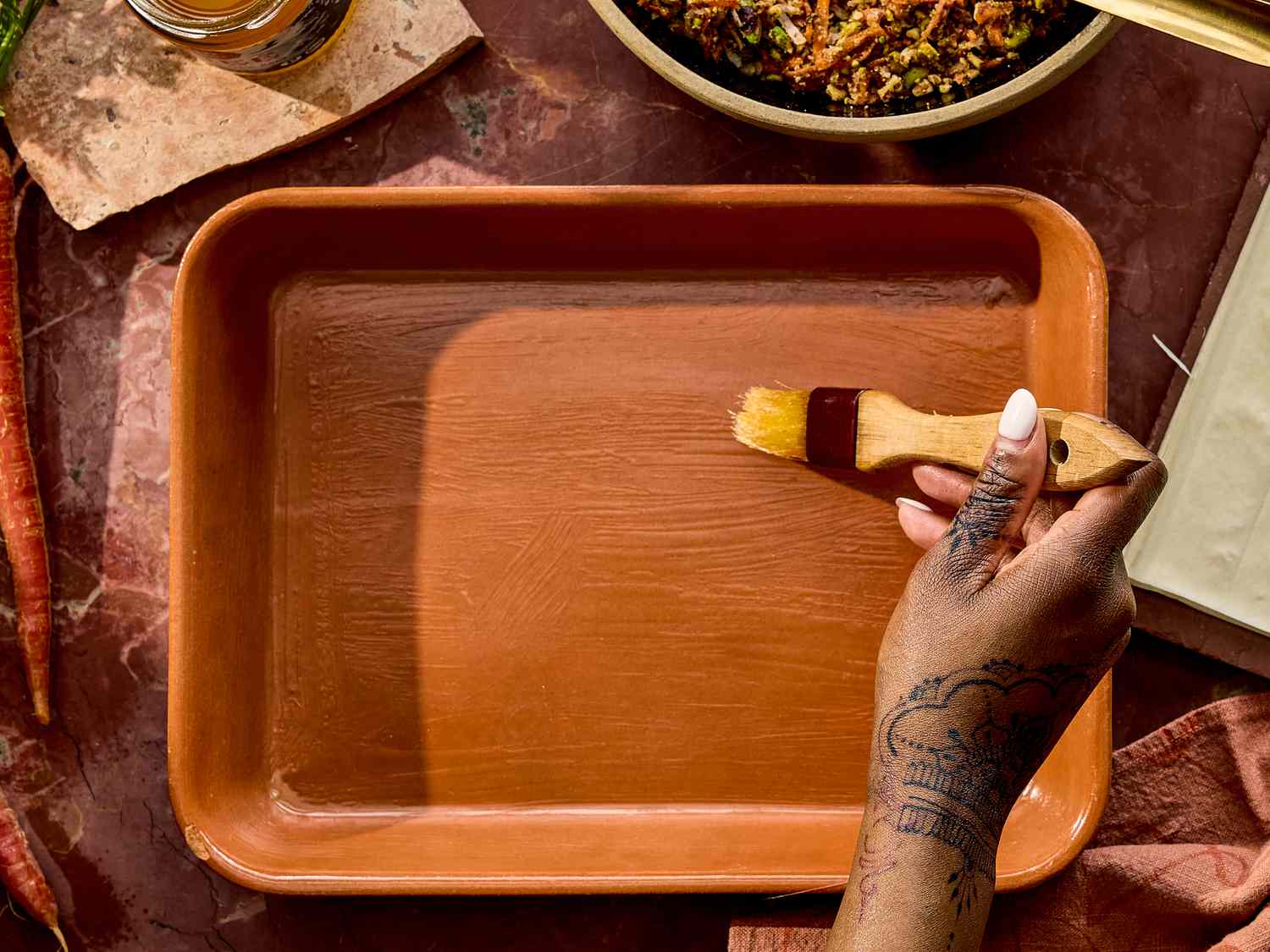 A person brushing the inside of a ceramic baking dish with oil, surrounded by prepared ingredients for carrot cake baklava