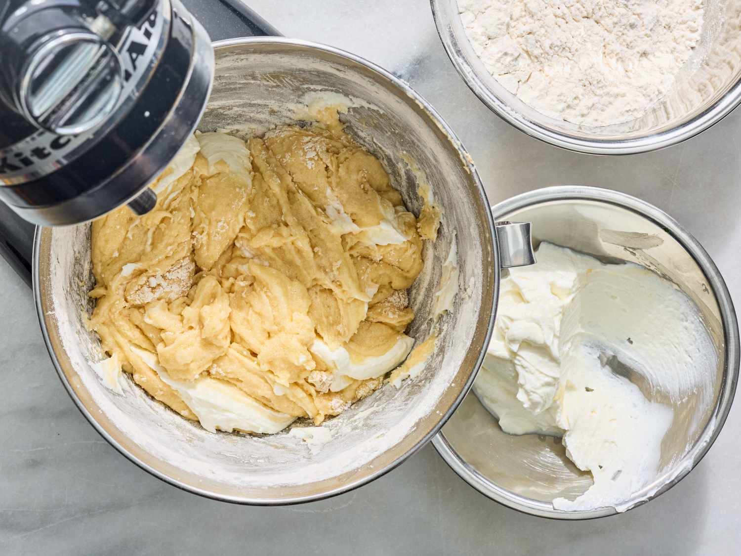 Mixing cake batter in a stand mixer with bowls of dry ingredients and cream nearby