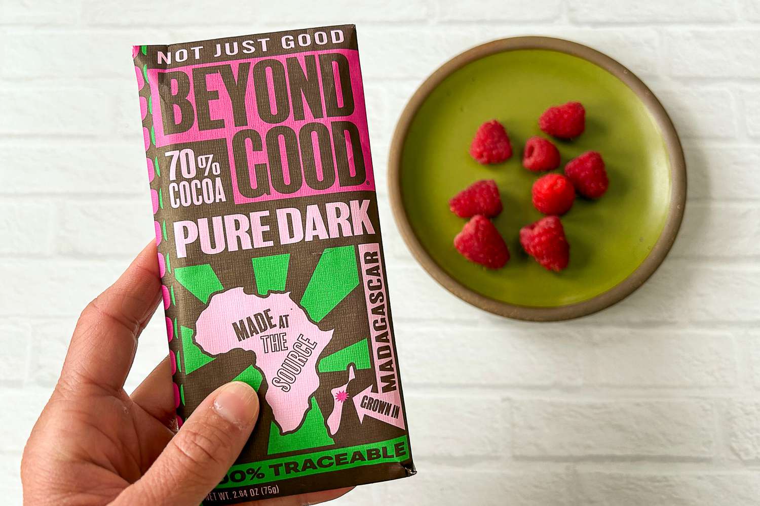 A person holding the Beyond Good Pure Dark 70% Bar next to a plate of raspberries