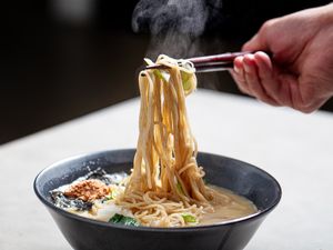 A hand lifting noodles with chopsticks from a bowl of ramen