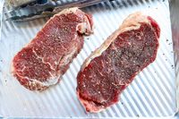 Two seasoned Butcherbox raw steaks on a metal pan with tongs nearby
