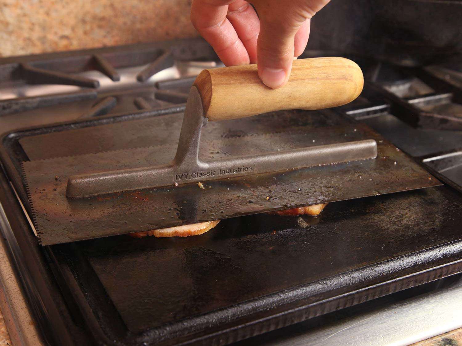 A finishing trowel is used to weight the slices of Canadian bacon as they brown on a griddle.