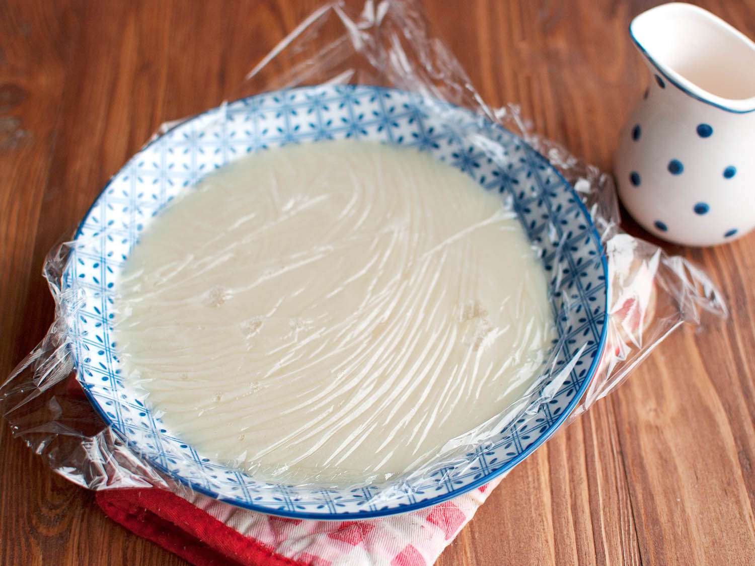 The pudding covered in plastic wrap as it rests in a bowl. 