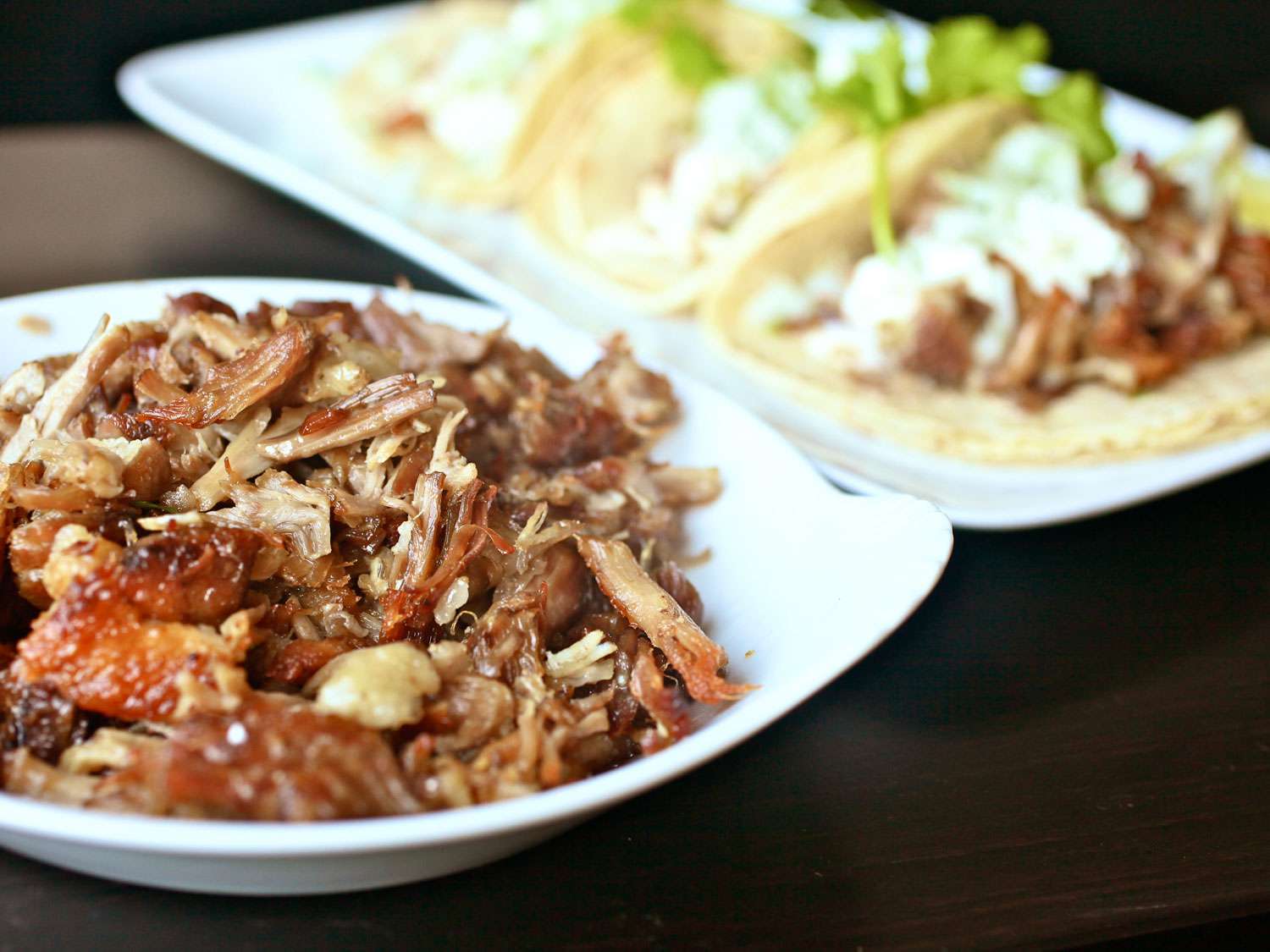 A bowl of no-waste carnitas served in a white bowl. A rectangular plate of dressed tacos is visible in the blurred background.