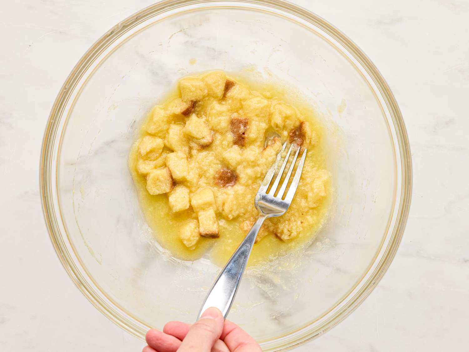 A hand holding a fork mashing ingredients in a clear glass bowl on a light surface