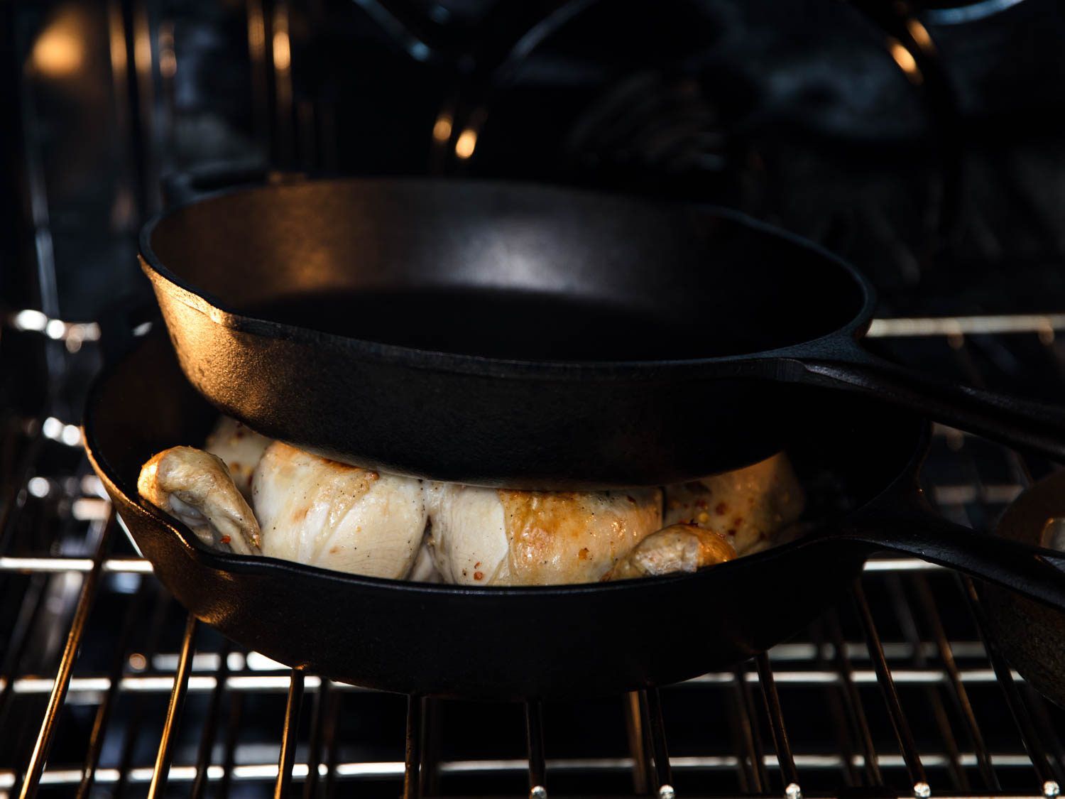 A cast iron skillet pressing down on a spatchcocked chicken set in another cast iron skillet, resting on an oven rack.
