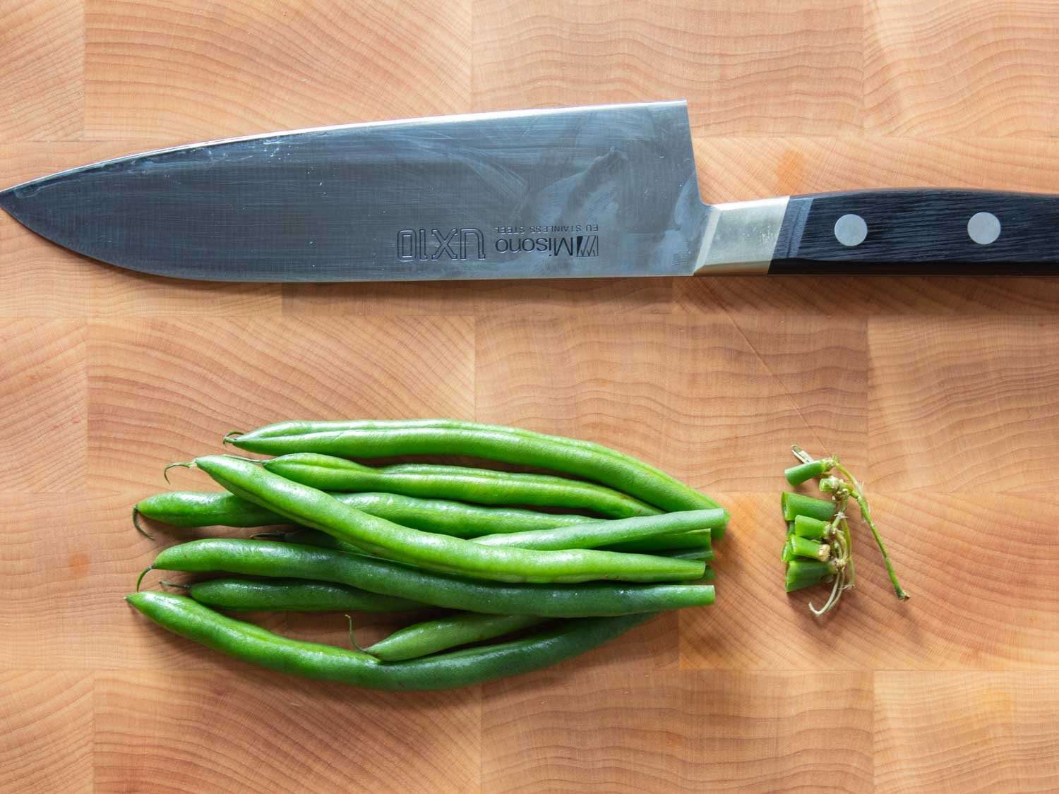 Closeup of green beans trimmed at the stem end with a santoku knife next them on a cutting board.