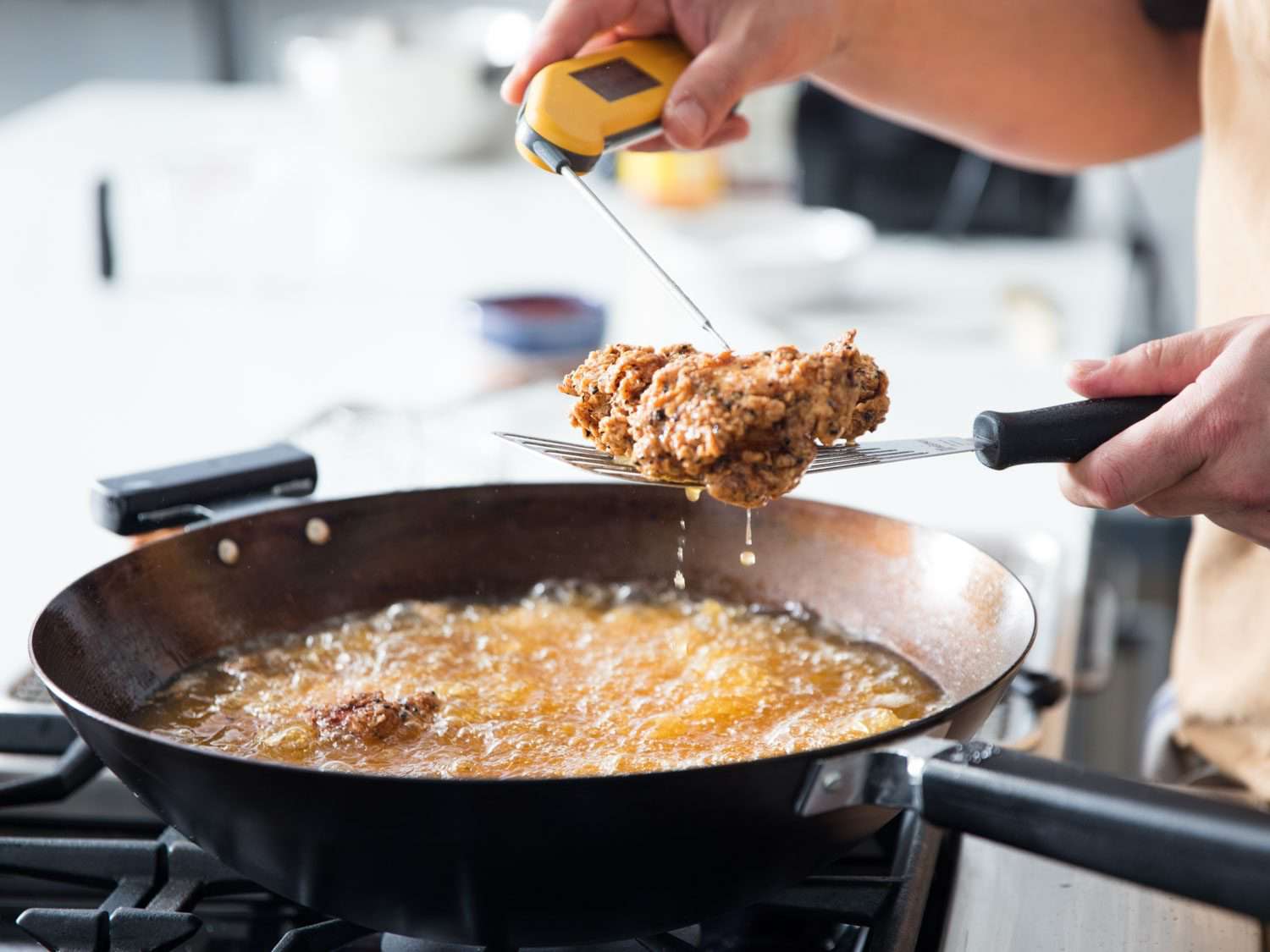 Digital thermometer being used to test the doneness of a piece of fried chicken, held aloft over a wok filled with bubbling oil and more pieces of frying chicken.