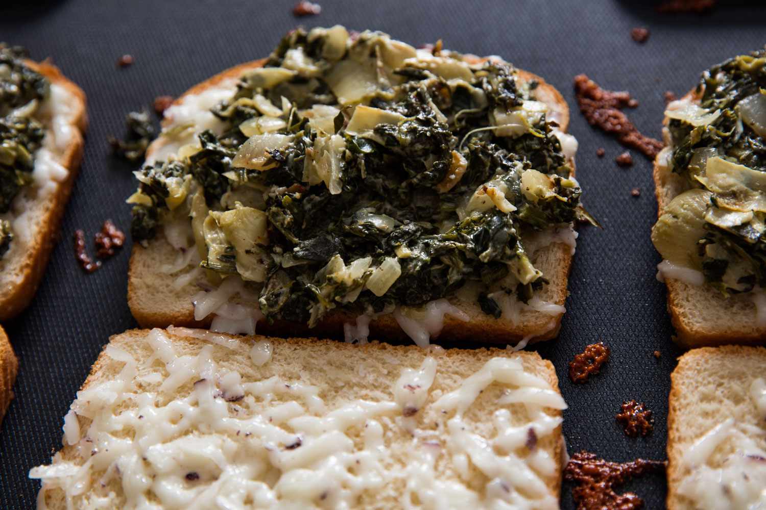 Closeup of the baked, cheese-covered bread slices. Half of the slices are covered with spinach-artichoke mixture.