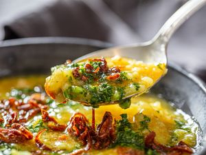 Closeup of a spoon lifting a portion of a dish with cooked rice garnished with herbs and fried shallots from a bowl
