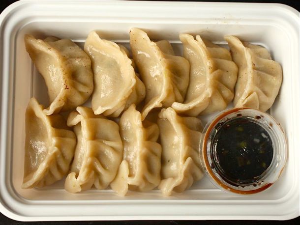 Homemade pork and cabbage dumplings with homemade wrappers, placed in a cardboard take-out container to replicate how they're served in a restaurant. There is a small plastic container of dipping sauce in the bottom right corner of the container.