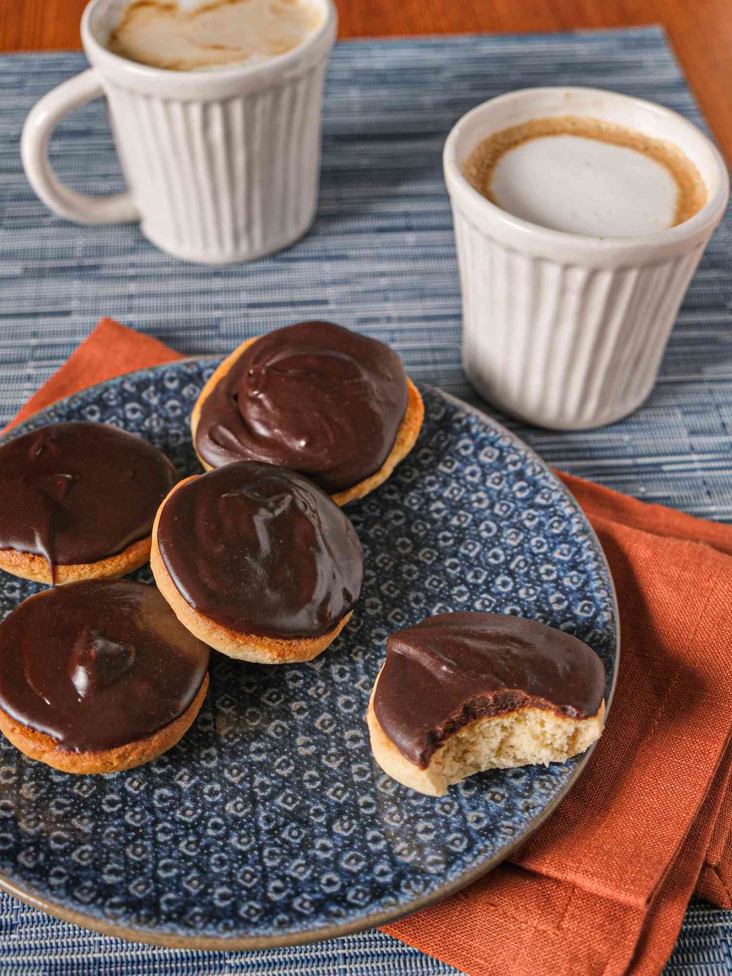 Baltimore Berger cookies on a plate, one with a bite taken out of it. Plate is on a red napkin, blue tablecloth and has 2 white cups of coffee 