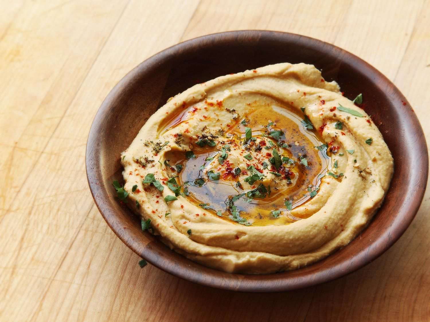 A wooden bowl of hummus drizzled with olive oil and garnished with herbs and paprika