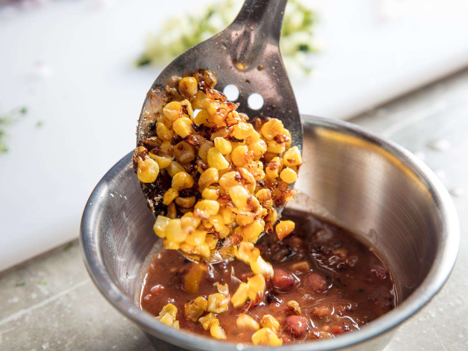 Charred corn kernels is added to a bowl of salsa with a slotted spoon.