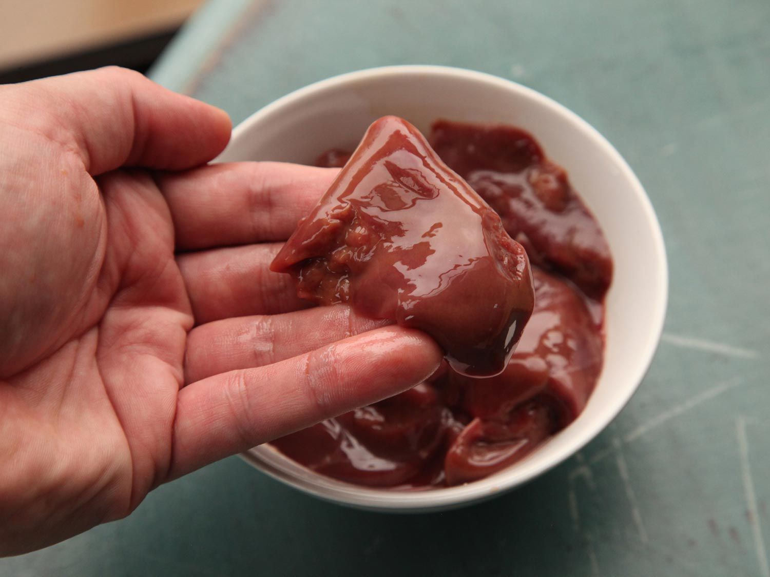 A hand holding a piece of liver over a bowl filled with cuts of liver.