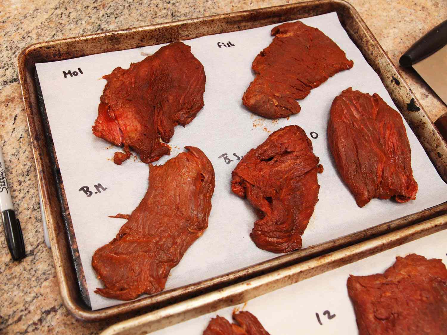 Steaks marinated using various flavor enhancers on parchment-lined baking sheets before grilling for carne asada.
