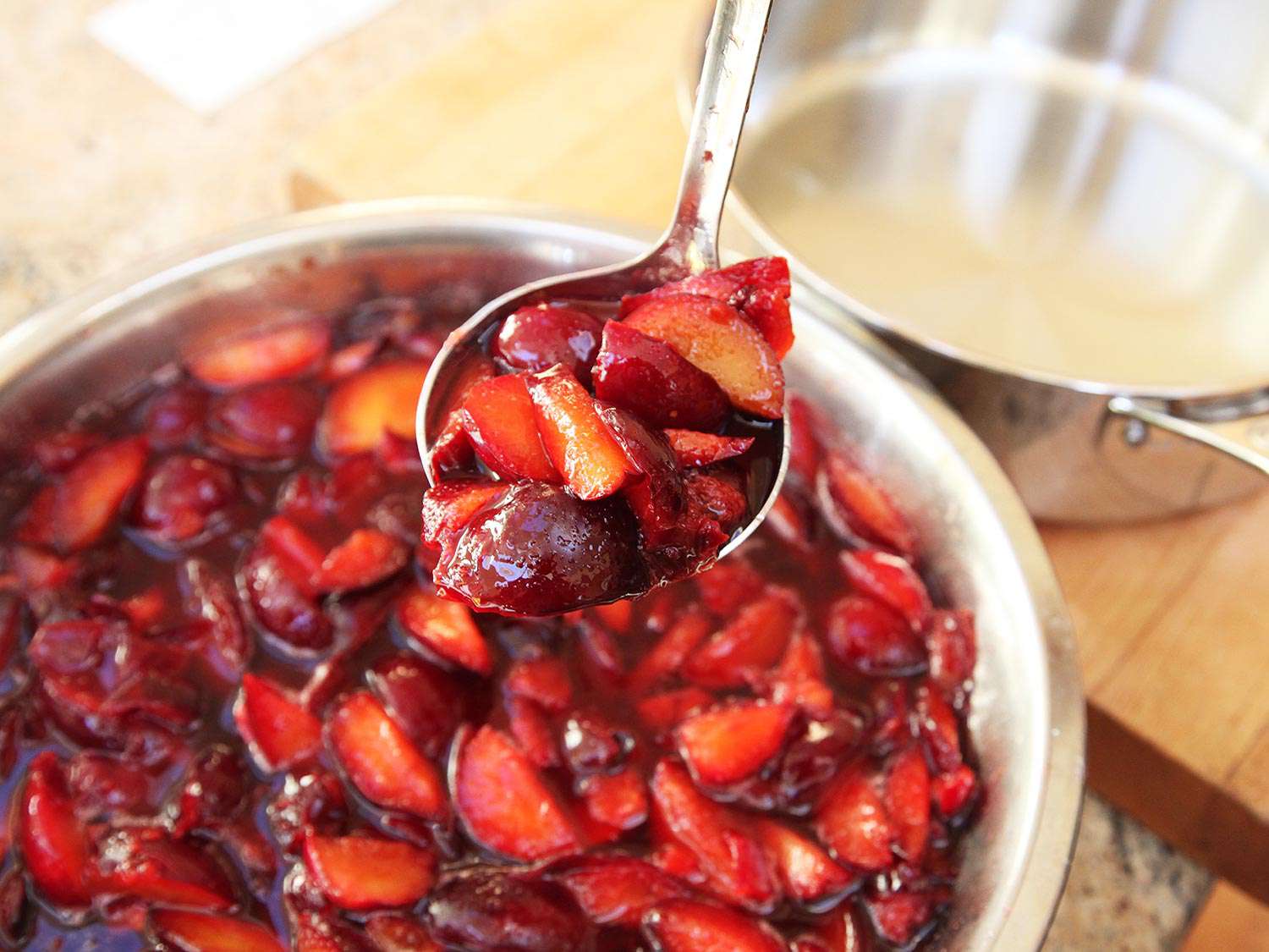 Close-up of a large spoon lifting a spoonful of macerated sliced plums from a large bowl.