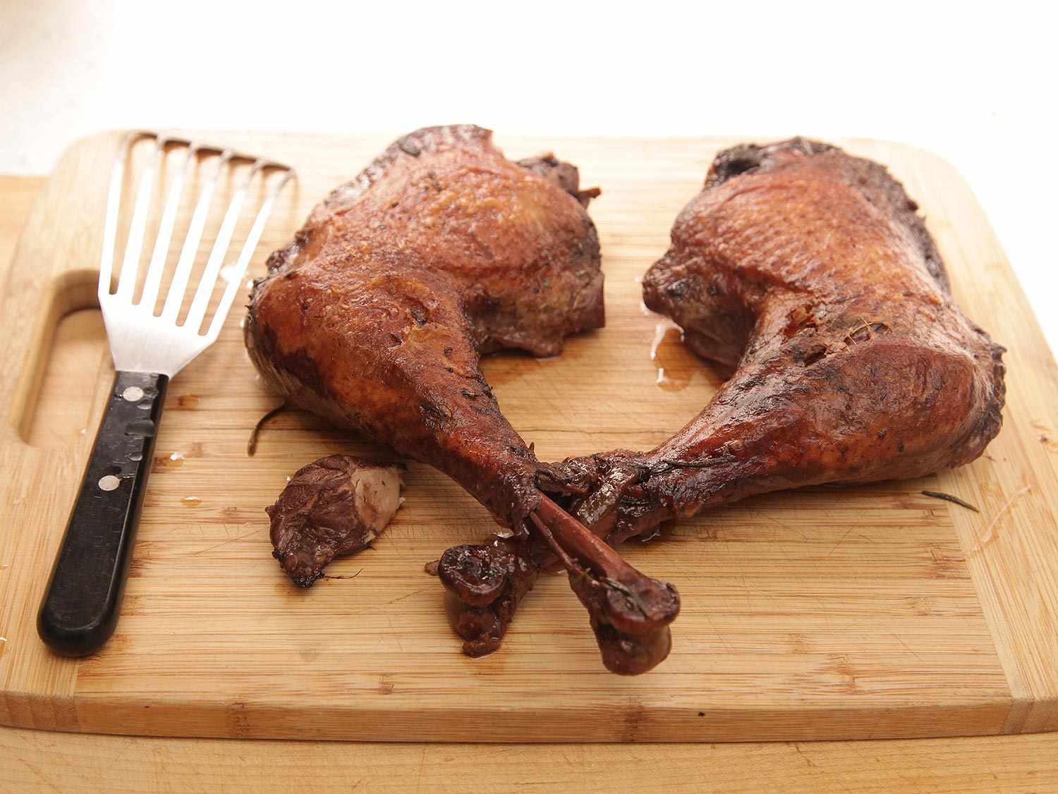 Two braised turkey legs resting on a cutting board next to a slotted spatula. 