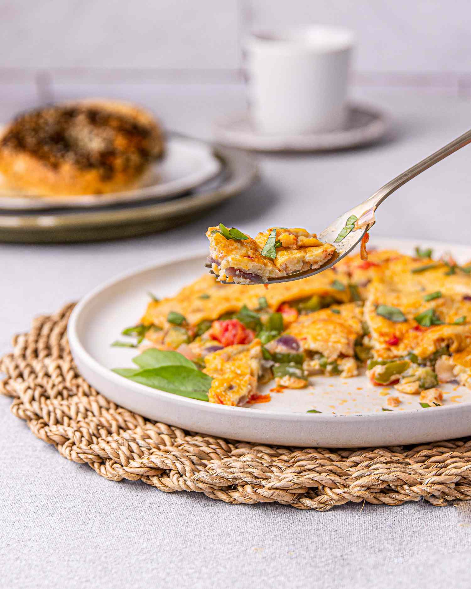 A plate with pieces of a Nigerian omelet a fork holding a bite a bagel and a coffee cup in the background