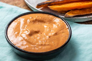 A bowl of chipotle mayonnaise on a light blue linen, with a plate of roasted vegetables behind it.