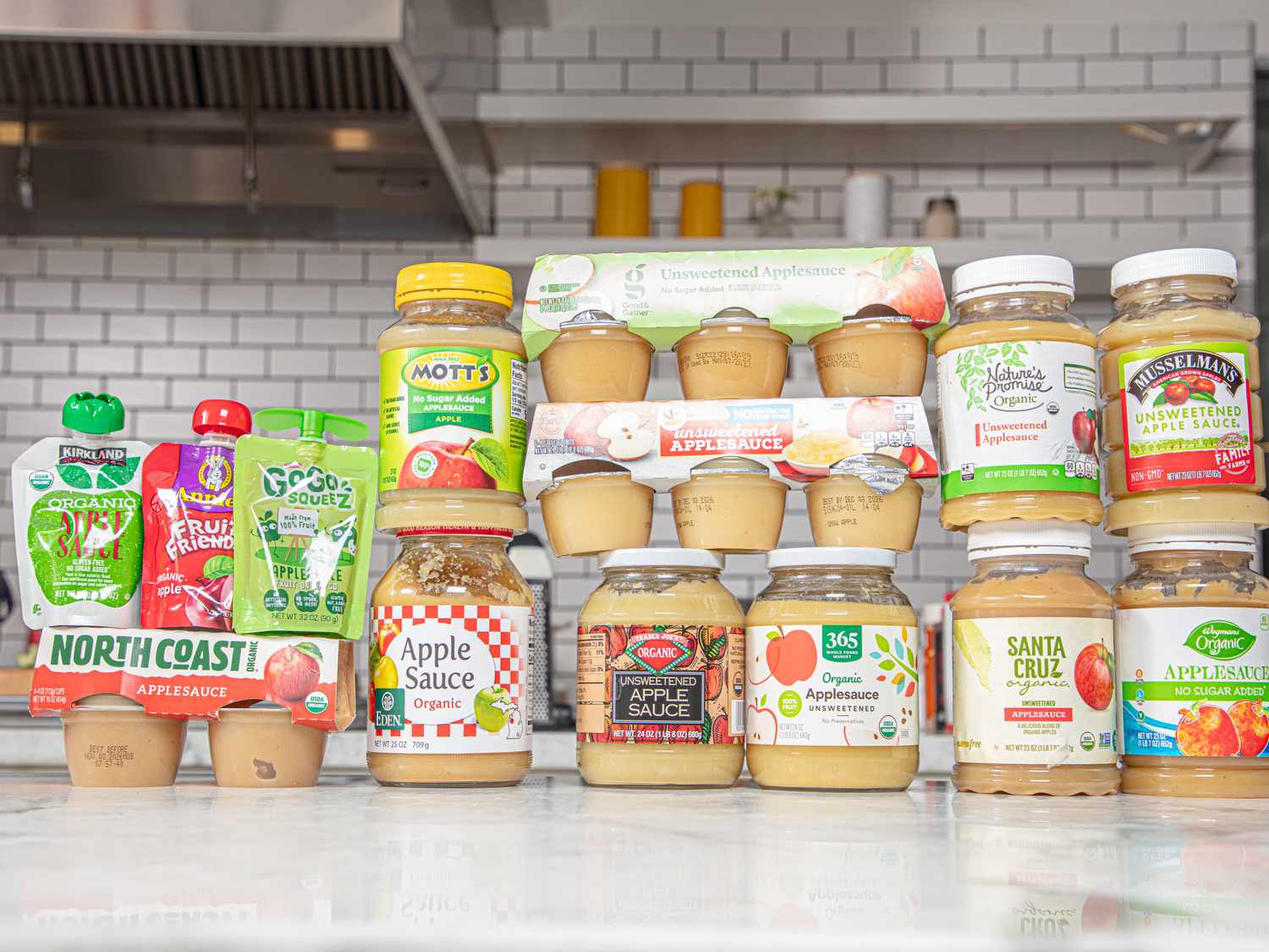 Various brands of applesauce in jars pouches and cups arranged on a counter in a kitchen setting
