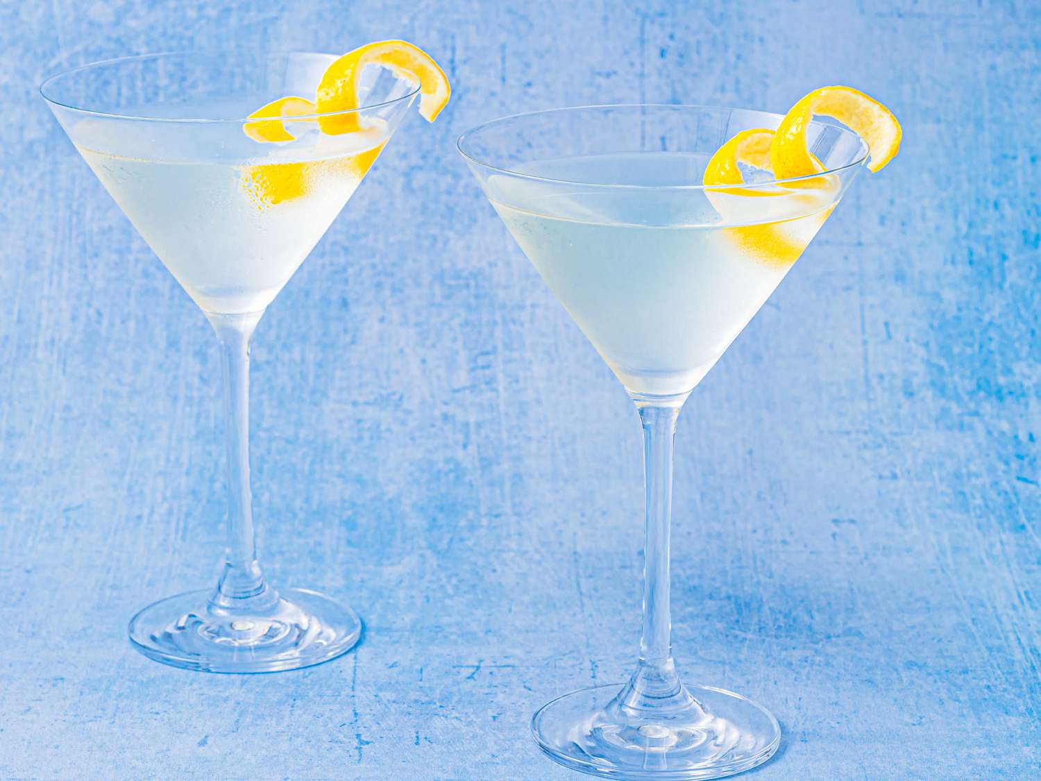 Two Martinis served with lemon twists on a distressed light blue surface.