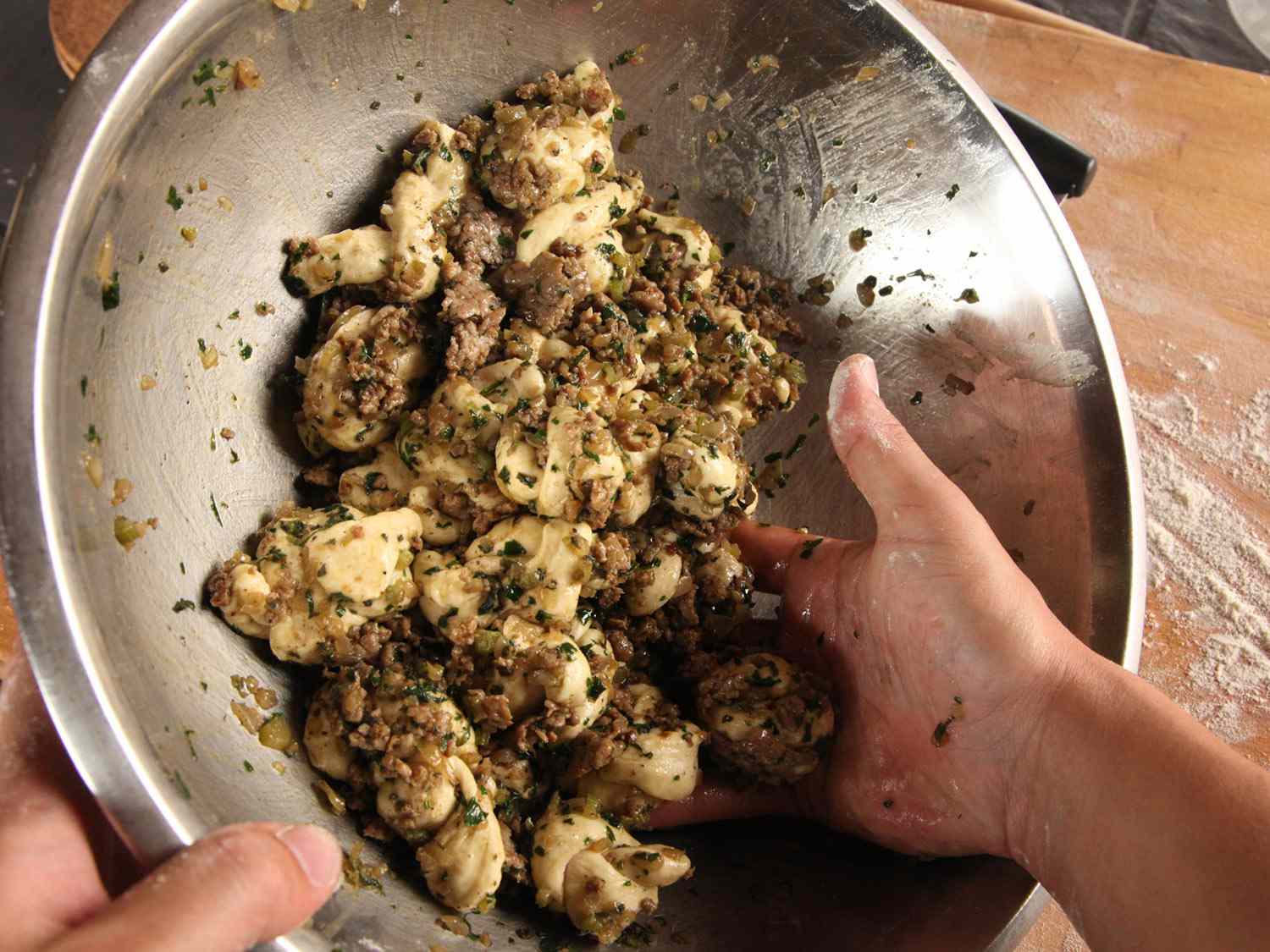 Using hands to toss knotted dough strips with sausage/aromatic mixture in a metal bowl