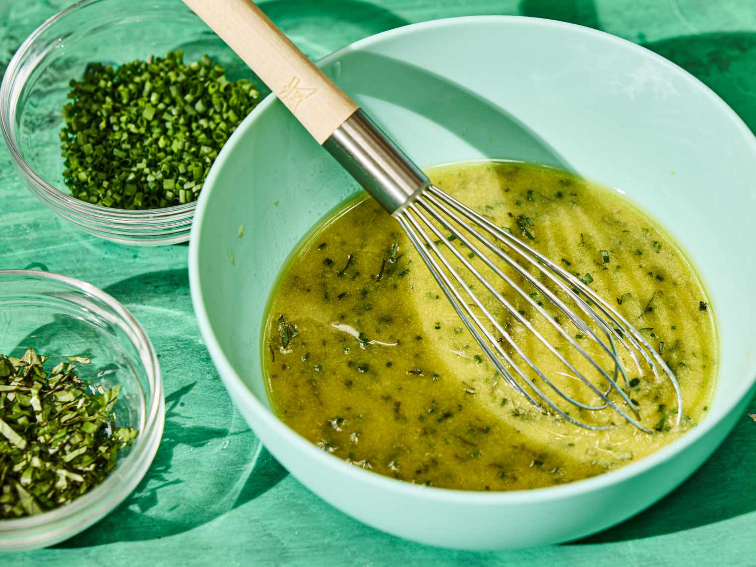 A whisk in a bowl with herb-infused liquid, surrounded by bowls of finely chopped herbs on a green surface