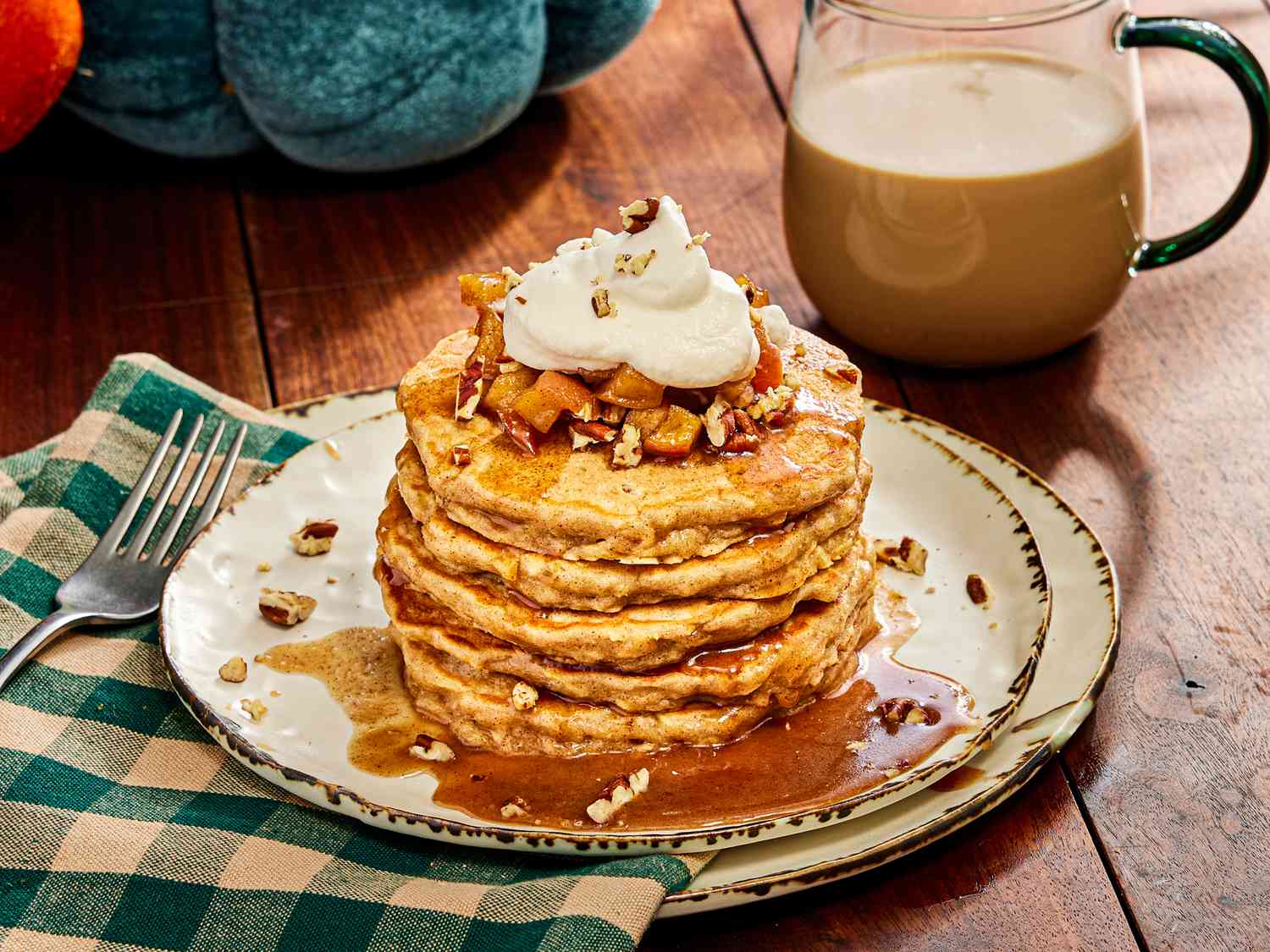 Stack of apple pie pancakes on white rustic plates, a green checked napkin, and warm drink