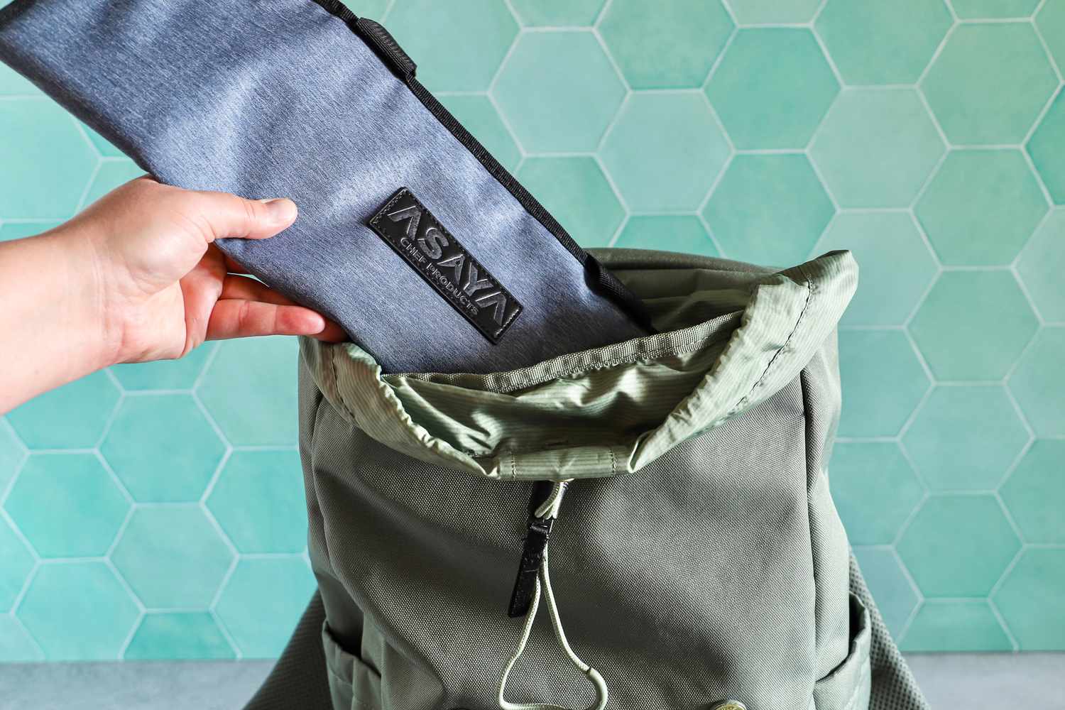 A hand placing a Asaya Chef Knife Roll Bag into an open backpack